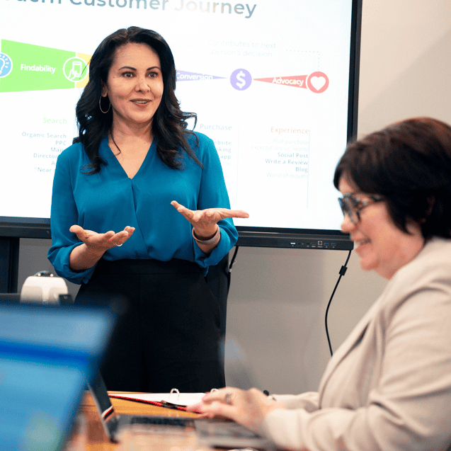A woman in a blue blouse stands and gestures while presenting a marketing strategy on a screen. Another woman in glasses and a beige blazer, likely her partner, sits at the table with laptops in the foreground.