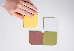 A hand places a yellow, rounded-edge square tile—symbolizing marketing—next to three other tiles (white, brown, and green) arranged together on a white surface, suggesting a thoughtful strategy.