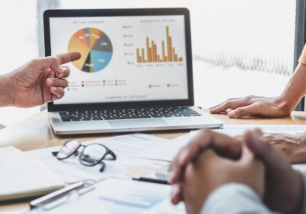 Three people meet at a table with documents and glasses, reviewing a marketing strategy on a laptop displaying a pie chart and bar graph. One partner points at the screen while the others listen and observe.