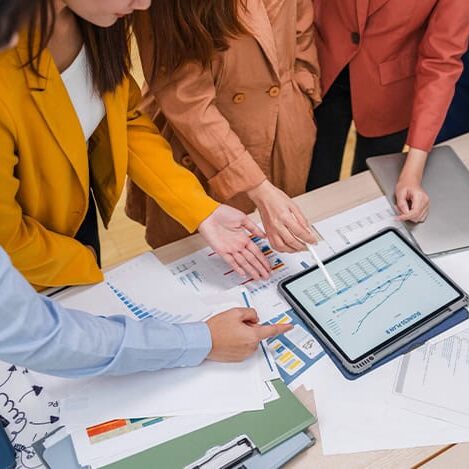 Four people in business attire stand around a desk covered with documents and charts, discussing a marketing graph displayed on a tablet. One partner points to the graph with a pen, highlighting their collaborative analysis and strategy development.