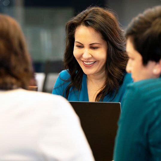 A woman with dark hair, wearing a teal top, smiles while collaborating on marketing strategy with two partners in a professional setting.