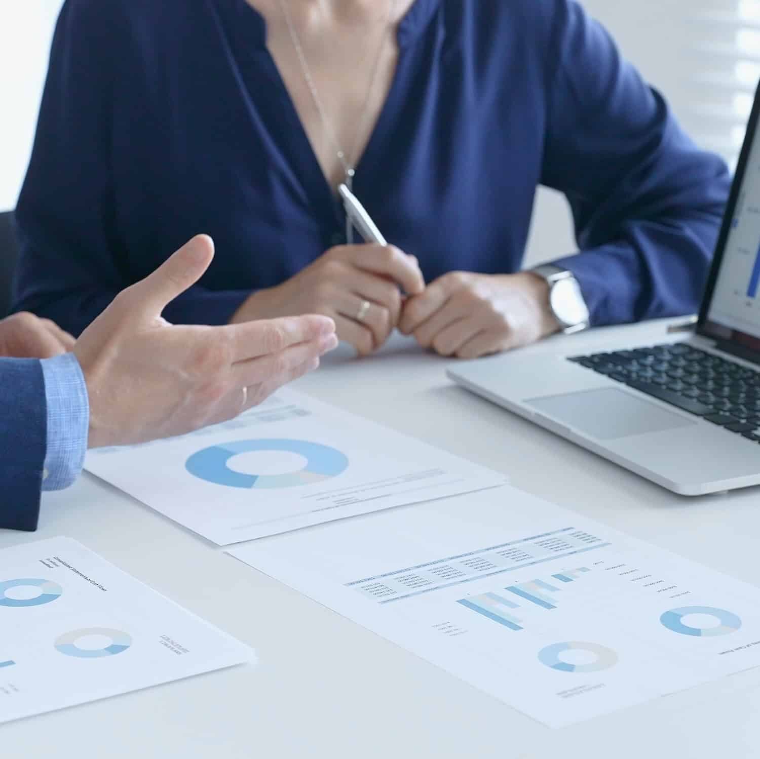 Two people sit at a desk reviewing printed charts and graphs, with a laptop displaying a graph. One person gestures while the other holds a pen, suggesting a discussion about marketing strategy or business partnership.