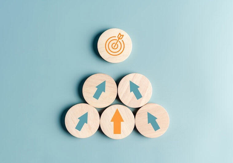 Five wooden circles on a blue background form a pyramid, symbolizing marketing strategy. Four lower circles have blue arrows pointing upward; the top circle has an orange target. The middle bottom circle’s arrow is orange, highlighting the key partner.
