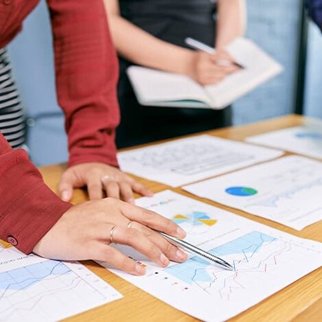 Three people stand at a table reviewing printed charts and graphs. One person points at a line graph with a pen, guiding their partners as they discuss marketing strategy while another takes notes in a notebook. The focus is on their hands and the documents.